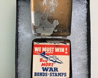 May include: Vintage metal match safe with a worn, tarnished finish. The open safe reveals a matchbox with the text "WE MUST WIN!" and "Buy MORE WAR BONDS * STAMPS" with an illustration of a plane. The match safe is in a black box with white padding.