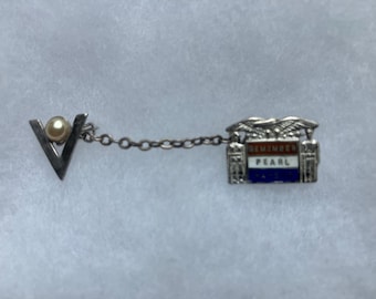 May include: A vintage pin with a silver-tone V-shaped design, featuring a pearl accent and a chain connecting to a rectangular emblem. The emblem reads "REMEMBER PEARL" in red, white, and blue.