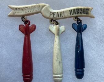 May include: Vintage brooch featuring a cream-colored banner with the words "REMEMBER PEARL HARBOR." Three dangling bomb-shaped charms are painted red, white, and blue. The brooch is a patriotic commemorative item.