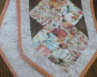 May include: Quilted table runner featuring a fall harvest theme. The center design showcases pumpkins and leaves in shades of orange, white, and green. The runner is bordered with a light orange polka dot trim and a quilted pattern.