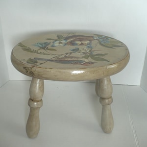 May include: A small, round wooden stool with a light beige, distressed finish. The top surface features a painted illustration of a bird perched on a branch near a nest with eggs, surrounded by blue and white flowers and green foliage.