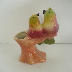 May include: A ceramic planter shaped like a tree stump with two birds perched on a branch. The birds are colored in shades of yellow, pink, and green with black beaks and eyes. The tree stump base is a light peach color with a glossy finish.