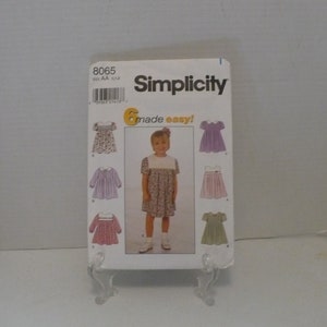 May include: Simplicity sewing pattern number 8065 for girls' dresses in sizes AA to 12. The pattern includes six different styles of dresses, including a floral print dress, a solid purple dress, a solid pink dress, and a solid green dress. The pattern is titled "6 made easy!"