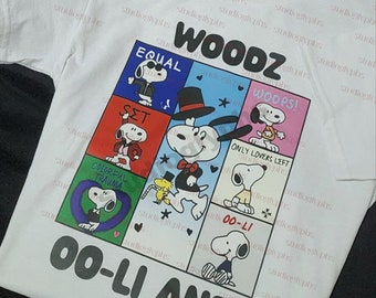May include: White t-shirt with a colorful graphic print of Snoopy characters in different poses. The text "WOODZ" is printed in black at the top of the graphic. The text "OO-LI AND" is printed in black at the bottom of the graphic.