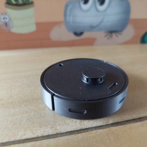 Könnte beinhalten: Ein schwarzes, kreisförmiges Gerät mit einem erhabenen Knopf in der Mitte und einer strukturierten Oberfläche. Das Gerät hat einen kleinen Schlitz an der Seite und liegt auf einer hellen Holzoberfläche. Der Hintergrund zeigt eine Cartoon-Illustration.