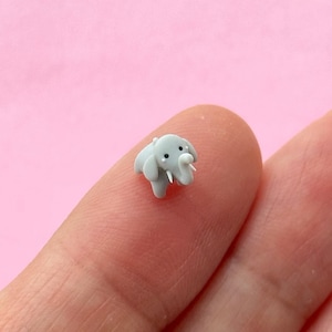 Könnte beinhalten: Eine winzige, graue Elefantenfigur mit schwarzen Augen und weißen Stoßzähnen. Der Miniatur-Elefant steht auf einem Finger vor einem rosa Hintergrund. Der Elefant ist eine kleine, detaillierte Skulptur.