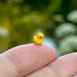 以下が含まれることがあります： オレンジ色のくちばしと黒い目を持つ小さな黄色のラバーダックが指先に座っています。 ミニチュアの鴨は明るい黄色で、背景はぼやけた緑色です。