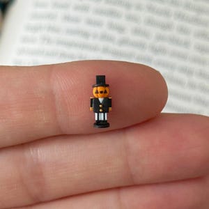May include: A miniature figurine shaped like a pumpkin-headed character wearing a black top hat and a black suit with white vertical stripes. The figure has a bright orange head with black triangular eyes and a small black mouth, standing upright.