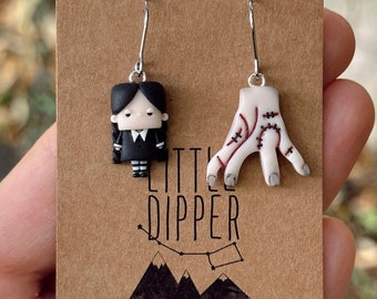 May include: A pair of dangle earrings featuring a black and white cartoon character and a white hand with red stitching. The earrings are displayed on a brown card with the text "LITTLE DIPPER" and contact information for the shop.