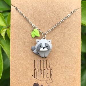 May include: A silver chain necklace with a small grey raccoon charm and two green leaves. The charm has black eyes and a black nose. The necklace is on a brown card with the text "LITTLE DIPPER" printed on it.