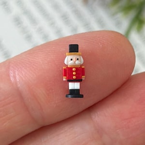 Könnte beinhalten: Eine Miniatur-Spielzeugsoldatenfigur mit roter Jacke, weißer Hose und schwarzem Hut. Die Figur hat goldene Akzente und steht auf einem schwarzen Sockel. Der Spielzeugsoldat ist eine klassische Weihnachtsdekoration.