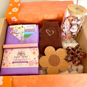 May include: A gift box containing a glass tumbler with a bamboo lid and autumn-themed decals, a pair of brown fuzzy socks with a white heart, a flower-shaped cork coaster, a leopard print hair claw, and a purple book-themed stationery set.