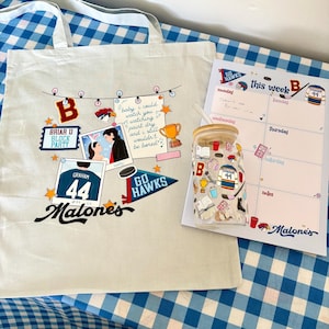 May include: A beige canvas tote bag featuring a graphic print of polaroids, a trophy, and text including 'Briar U Block Party' and 'Malones'. Next to it is a glass tumbler with a bamboo lid and a weekly planner pad with 'Go Hawks' and 'Malones' text.