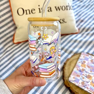 May include: A clear glass tumbler with a bamboo lid and a glass straw, decorated with colourful illustrations of books, flowers, hearts, and a cartoon character reading. The background includes a pillow with text and a striped pattern.