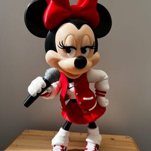 May include: A Minnie Mouse figurine wearing a red and white varsity jacket, a red pleated skirt, and red and white trainers. The character has a large red bow on her head, holds a grey microphone in one hand, and features black ears.