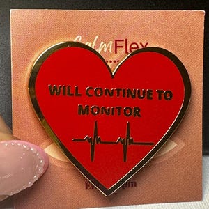 May include: A heart-shaped enamel pin with a gold border. The pin is red with the words "WILL CONTINUE TO MONITOR" and a heartbeat graphic. The pin is on a peach-colored card with the text "Calm Flex".
