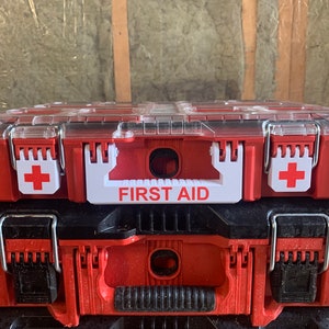 Puede incluir: Botiquín rojo y negro con tapa transparente. El kit tiene un asa blanca con las palabras "FIRST AID" en rojo. Una cruz blanca está en el lateral del kit. El kit está apilado encima de otro kit negro y rojo.