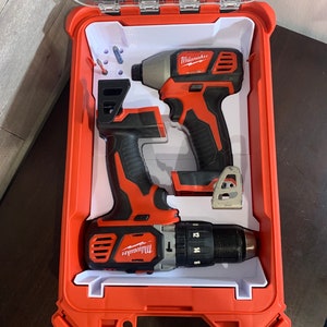 Peut inclure: Deux outils électriques Milwaukee, une perceuse et un tournevis, sont rangés dans un coffret rigide en plastique rouge et blanc. Le coffret est doté d'un couvercle transparent et d'un insert en mousse blanche pour maintenir les outils en place.