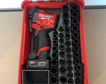 Op de afbeelding: Rode Milwaukee FUEL slagmoersleutel met een zwarte en grijze batterij en een koffer vol zwarte bussen. De koffer is rood met een doorzichtig deksel.