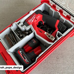 Op de afbeelding: Een rode en zwarte Milwaukee FUEL boormachine met twee rode lithium-ion batterijen, een grijze organizer met verschillende boorbits en een doorzichtig plastic deksel. De boormachine bevindt zich in een rode gereedschapskoffer.