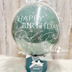 May include: A teal balloon with silver confetti and the words "HAPPY BIRTHDAY Dad" in white script. The balloon is presented on a teal box with a tag saying "HAPPY 64TH PULL ME!" and tied with a white ribbon. The Blue Sloth logo is visible.