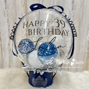 May include: A clear balloon with the text "HAPPY 39 BIRTHDAY Salem" contains blue and white balloons and silver confetti. The balloon sits atop a navy blue box with the text "BEST HUSBAND AND FATHER". A navy blue ribbon and bow adorn the top.