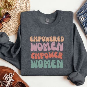 May include: A dark gray sweatshirt with the text "EMPOWERED WOMEN EMPOWER WOMEN" in a retro, colorful font.