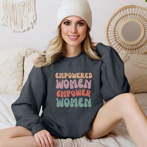 May include: A dark gray sweatshirt with the text "EMPOWERED WOMEN EMPOWER WOMEN" in colorful, wavy letters. The sweatshirt is being worn by a person with long blonde hair and a white beanie.