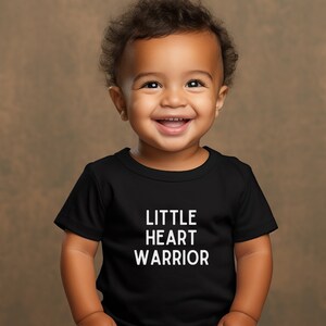 May include: A smiling baby wearing a black t-shirt with the words "LITTLE HEART WARRIOR" printed in white.
