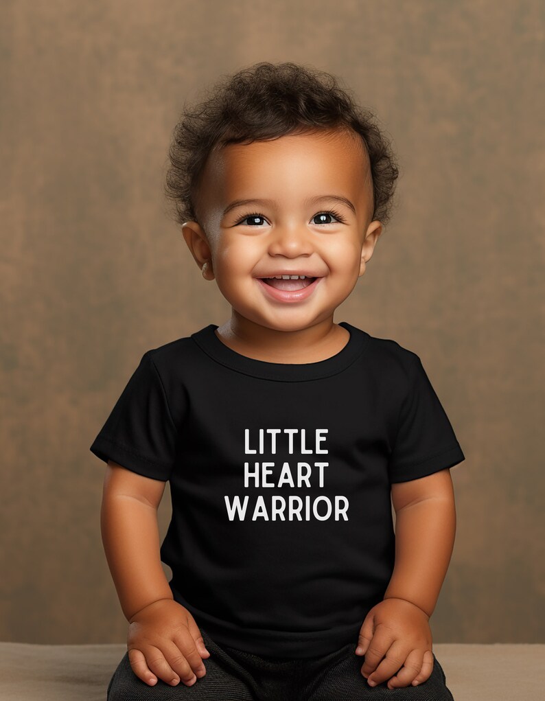 May include: A smiling baby wearing a black t-shirt with the words "LITTLE HEART WARRIOR" printed in white.