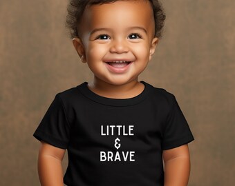 May include: A smiling toddler wearing a black t-shirt with the text "LITTLE & BRAVE" in white.