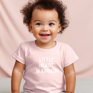 May include: A pink baby t-shirt with the text "LITTLE HEART WARRIOR" in white. A baby with curly brown hair is wearing the shirt and smiling.