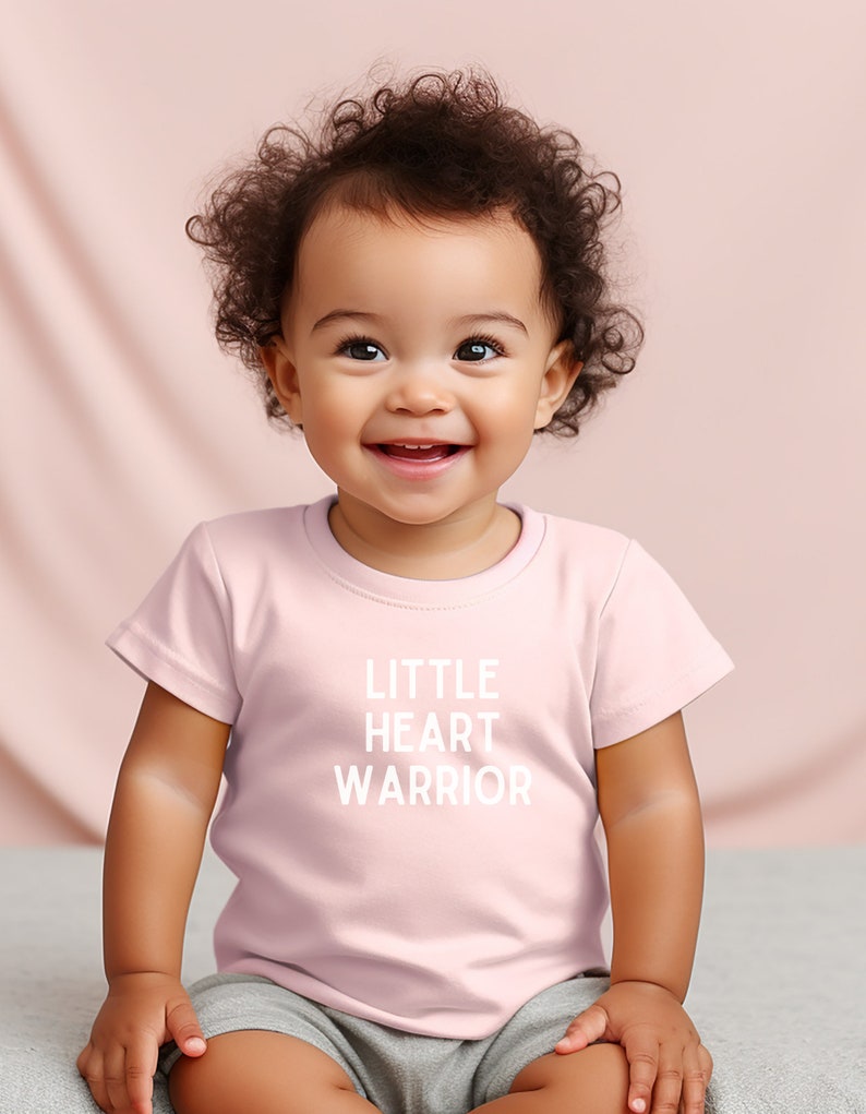 May include: A pink baby t-shirt with the text "LITTLE HEART WARRIOR" in white. A baby with curly brown hair is wearing the shirt and smiling.