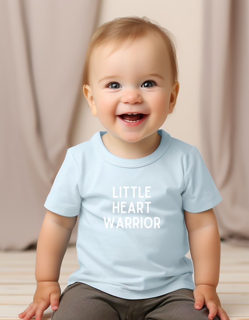 May include: A smiling baby wearing a light blue t-shirt with the text "LITTLE HEART WARRIOR" printed in white.