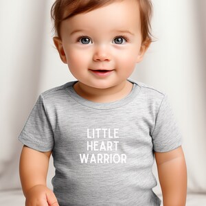May include: A gray short-sleeved t-shirt with the text "LITTLE HEART WARRIOR" printed in white on the front.