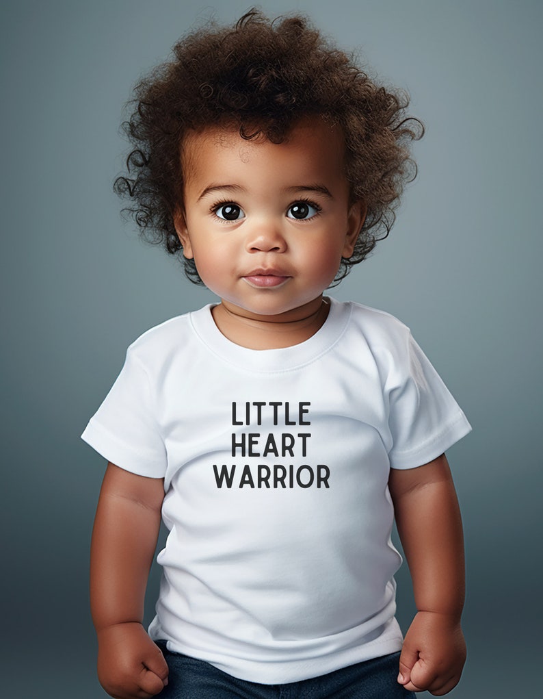 May include: A young child wearing a white t-shirt with the text "LITTLE HEART WARRIOR" printed on it. The child has curly brown hair and is looking directly at the camera.