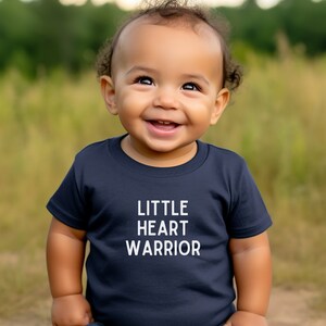May include: A young child wearing a navy blue t-shirt with the text "LITTLE HEART WARRIOR" in white. The child is smiling and looking up.