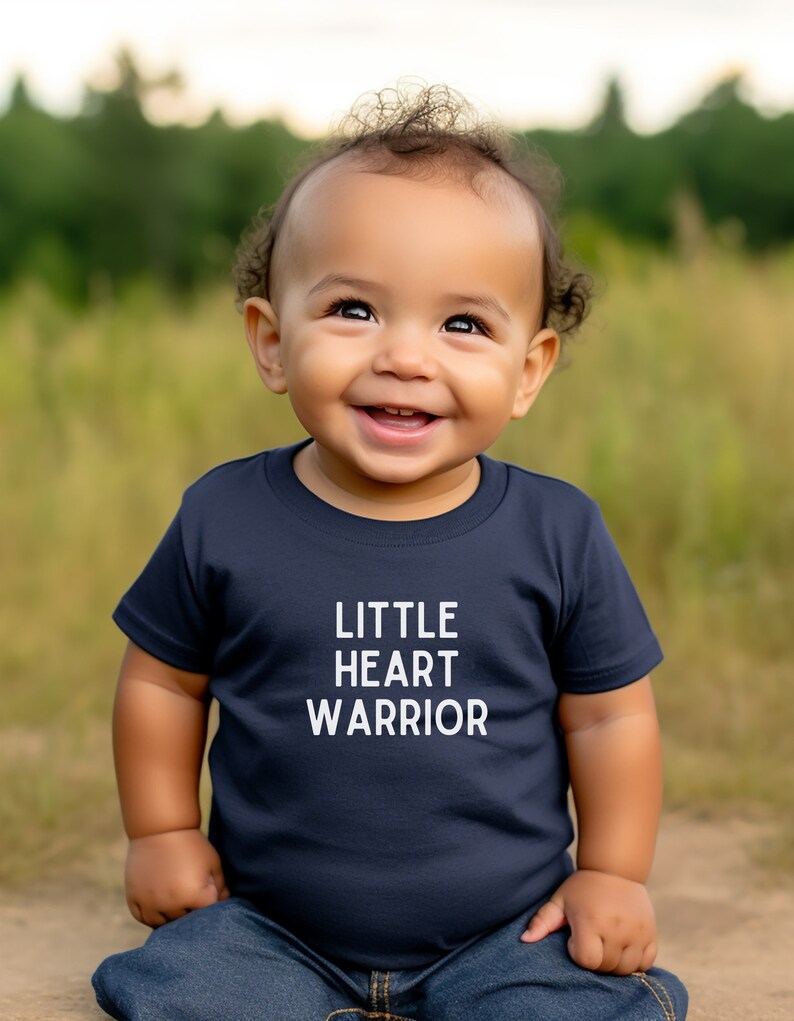 May include: A young child wearing a navy blue t-shirt with the text "LITTLE HEART WARRIOR" in white. The child is smiling and looking up.