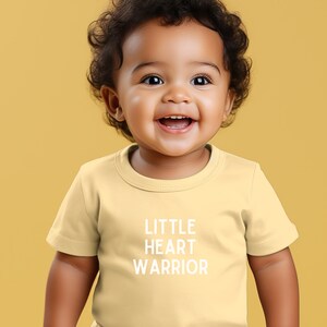 May include: A yellow short-sleeved t-shirt with the text "LITTLE HEART WARRIOR" printed in white on the front. A young child is wearing the shirt and smiling.