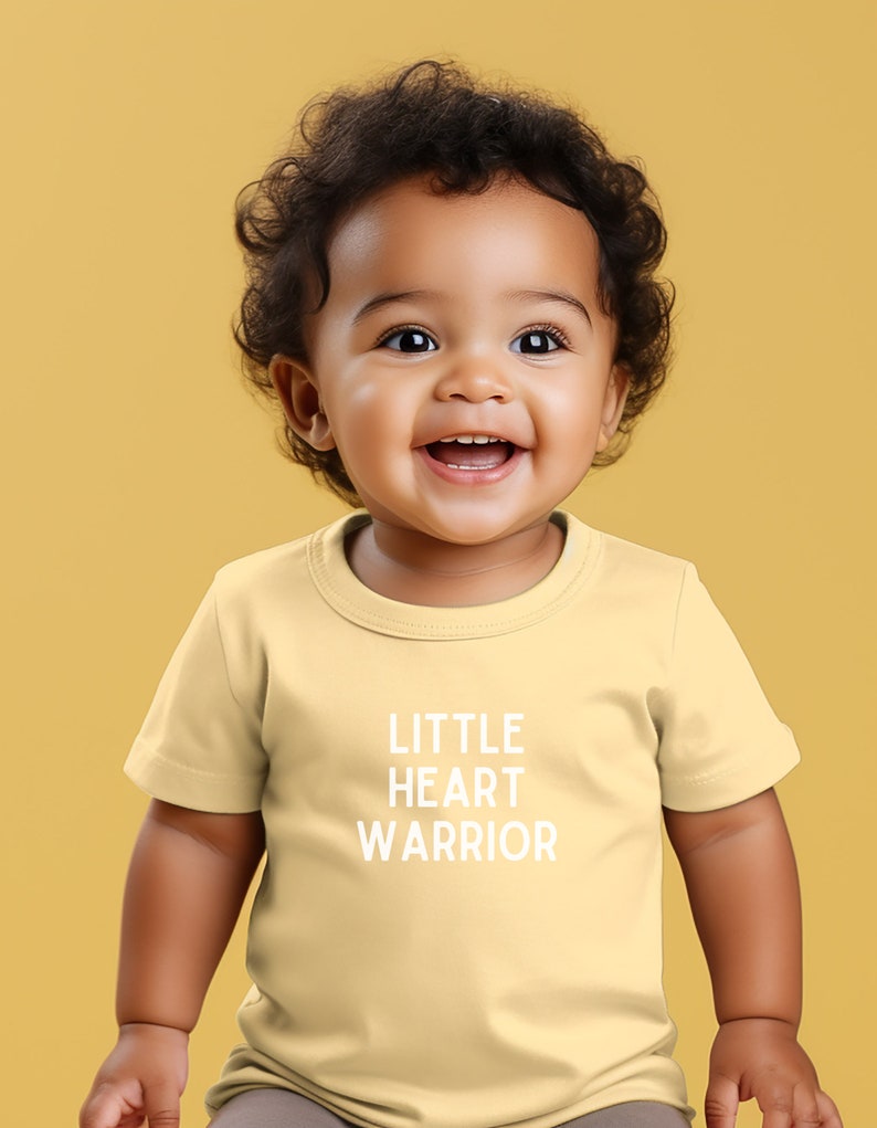 May include: A yellow short-sleeved t-shirt with the text "LITTLE HEART WARRIOR" printed in white on the front. A young child is wearing the shirt and smiling.