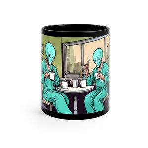 May include: A black coffee mug with a graphic of two green aliens sitting at a table with coffee cups. The aliens are wearing blue jumpsuits and have large heads and eyes.