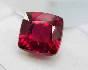 Puede incluir: Una gema facetada de talla cojín con un tono rojo intenso. La gema es cuadrada con esquinas redondeadas, reflejando la luz en sus múltiples facetas. La piedra se encuentra sobre un fondo blanco, resaltando su color y claridad.