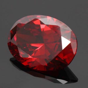 Puede incluir: Una gema facetada, de forma ovalada y color rojo intenso. La superficie de la gema refleja la luz, creando reflejos y sombras brillantes. La piedra se presenta sobre un fondo oscuro, resaltando su brillo. Esta gema podría usarse en joyería.