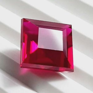 Può includere: Una gemma rosso rubino tagliata a forma quadrata e sfaccettata. La gemma è di un rosso intenso e saturo con un'apertura quadrata al centro. Le sfaccettature catturano la luce, creando riflessi e ombre. La gemma è su uno sfondo bianco a coste.