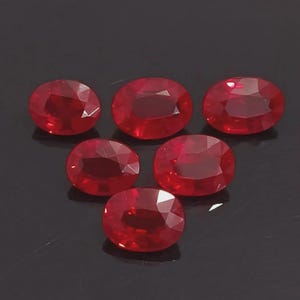 May include: Six oval-cut, faceted rubies. The gemstones are a deep, vibrant red colour and are arranged on a dark surface. The rubies have a polished, reflective surface, showcasing their internal brilliance.