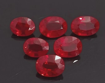 Puede incluir: Seis rubíes ovalados y facetados. Las piedras preciosas son de un color rojo intenso y vibrante y están dispuestas sobre una superficie oscura. Los rubíes tienen una superficie pulida y reflectante, mostrando su brillo interno.