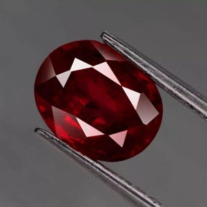 Pode incluir: Uma gema facetada, em forma oval, de cor vermelha escura, segurada por pinças prateadas. A gema tem múltiplas facetas que refletem a luz, criando reflexos brancos brilhantes contra a rica cor vermelha. O fundo é cinza liso.