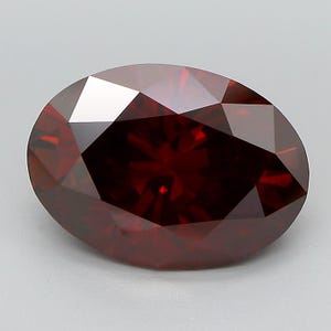 May include: A faceted, oval-shaped, deep red gemstone. The gem has a brilliant cut with multiple facets that reflect light, creating a sparkling effect. The color is a rich, dark red, with a hint of brown. The gemstone is set against a neutral background.