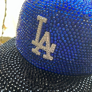 Puede incluir: Una gorra de béisbol azul y negra con un gran logotipo blanco "LA". La gorra está cubierta de diamantes de imitación.