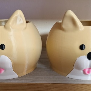 May include: Two ceramic dog-shaped planters. The planters are tan with black eyes, a black nose, white muzzle, and a pink tongue. The ears are upright. The planters are sitting on a wooden shelf.